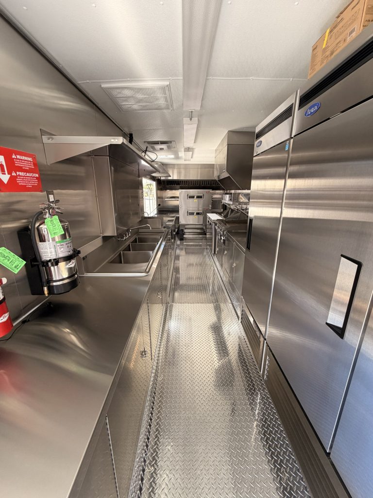 Commercial food trailer interior with stainless steel equipment, refrigeration, sinks, ventilation, and diamond-plate flooring.