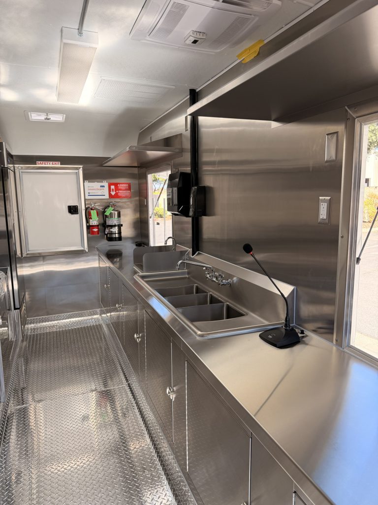 Custom food trailer interior with stainless steel sinks, fire suppression equipment, safety exit, storage, and commercial kitchen finishes.