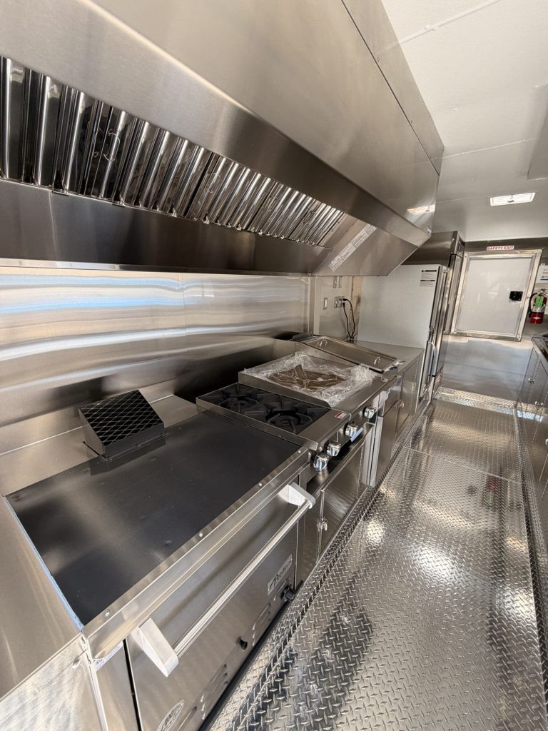 Custom food trailer cooking line with commercial ventilation hood, range equipment, stainless steel work surfaces, and diamond-plate flooring.
