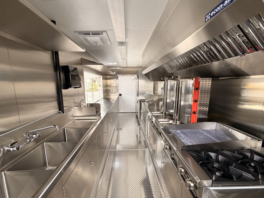 Stainless steel commercial food trailer interior with triple sink, flat top grill, shawarma machine, gas burners, and ventilation hood.