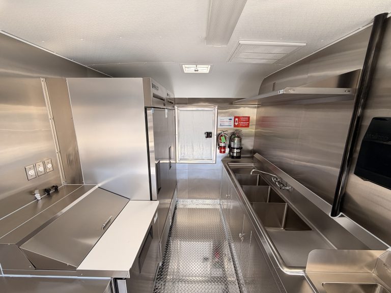 Smashed Broz Burgers food trailer full interior view with stainless steel prep counters, three-compartment sink, fire extinguisher, rear entry door and diamond plate flooring