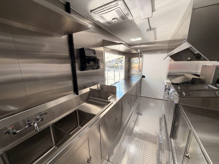 Shorter's BBQ and Catering food trailer sink zone with three-compartment sink, hand wash sink, paper towel dispenser and stainless steel prep shelf toward serving window