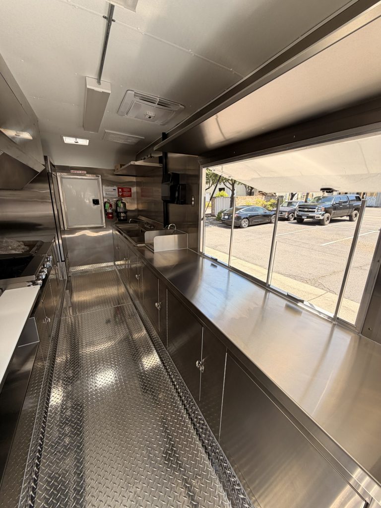 Custom food trailer interior with stainless steel counters, service window, sinks, storage cabinets, ventilation, and diamond-plate flooring.