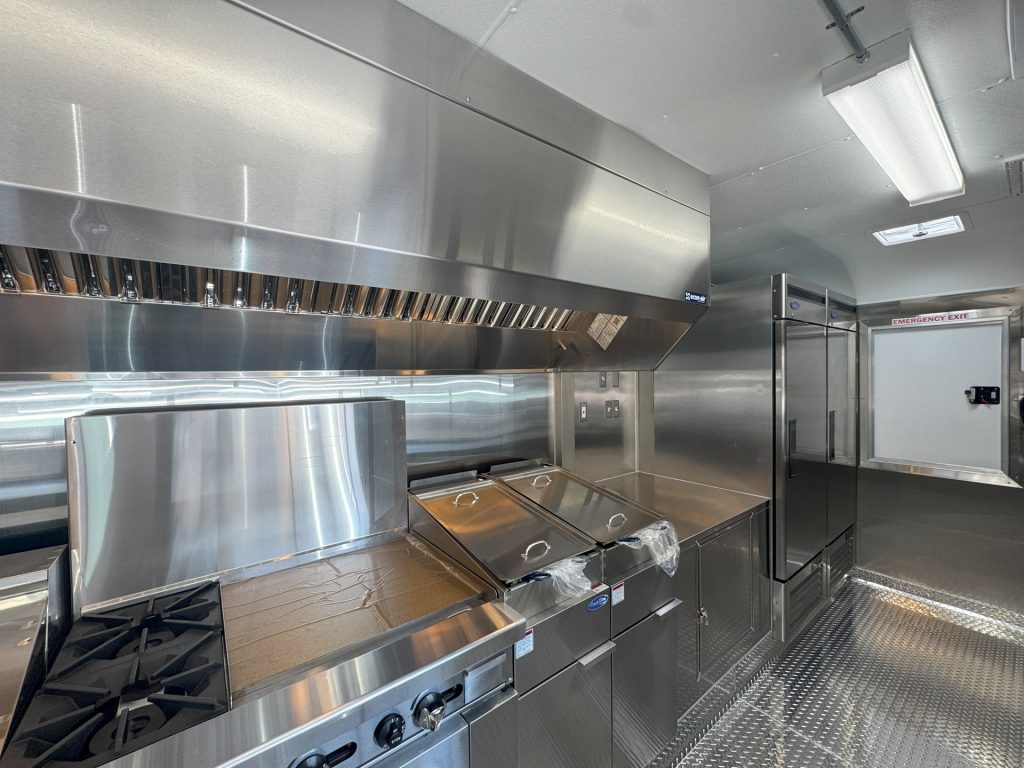 Commercial food trailer kitchen with stainless steel cooking equipment, exhaust hood, and diamond-plate flooring.
