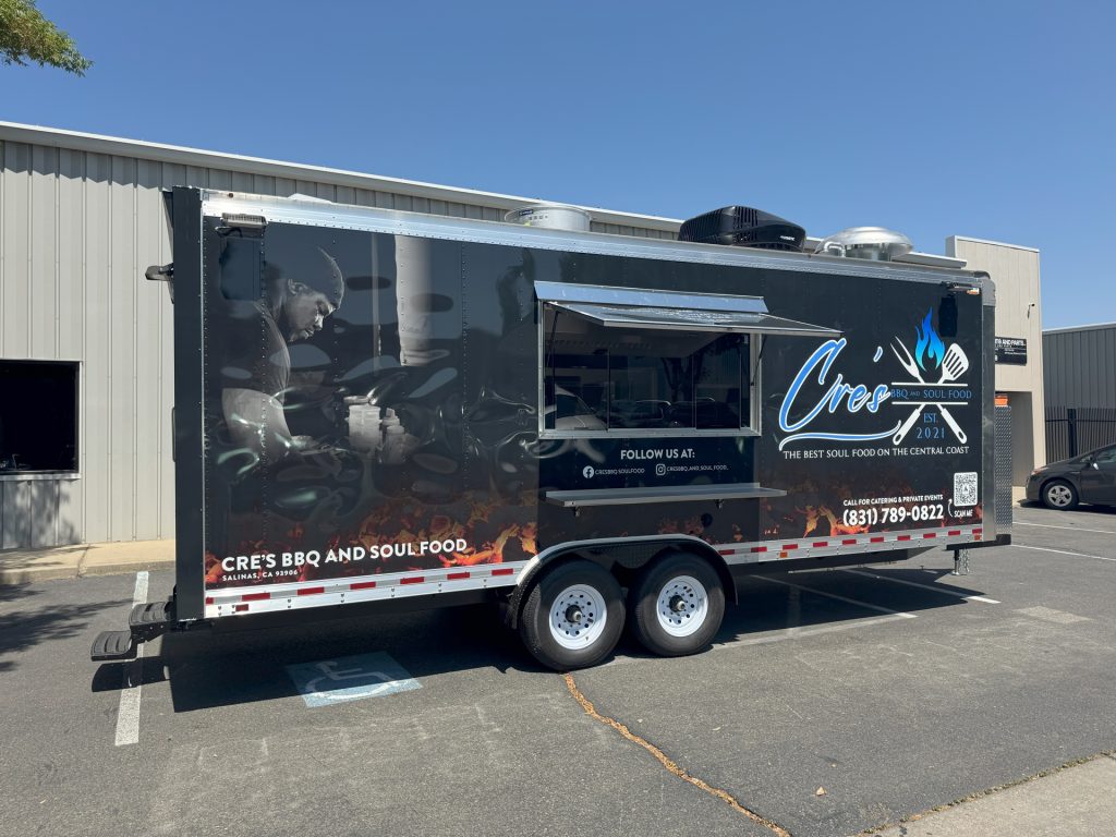 Black BBQ food trailer with blue graphics, serving window, and tandem axles.