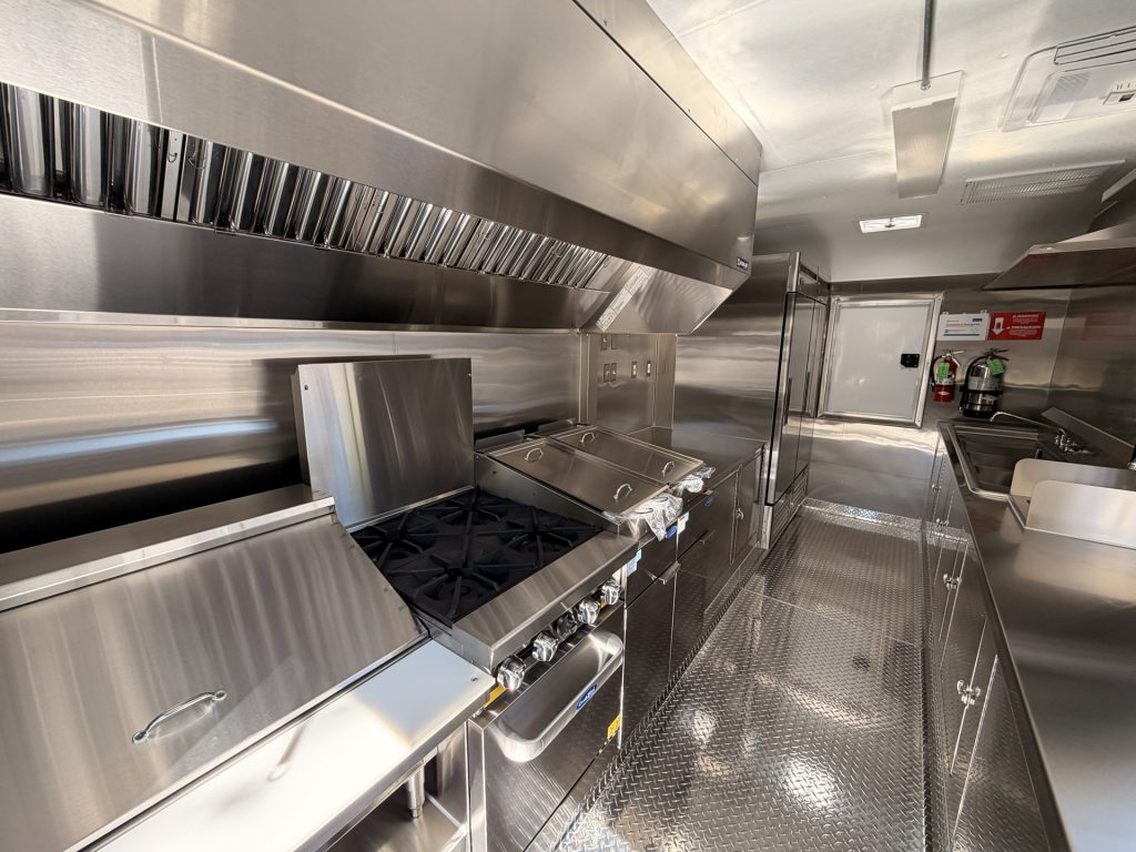 Commercial food trailer kitchen with gas cooking range, stainless steel exhaust hood, and preparation counters.