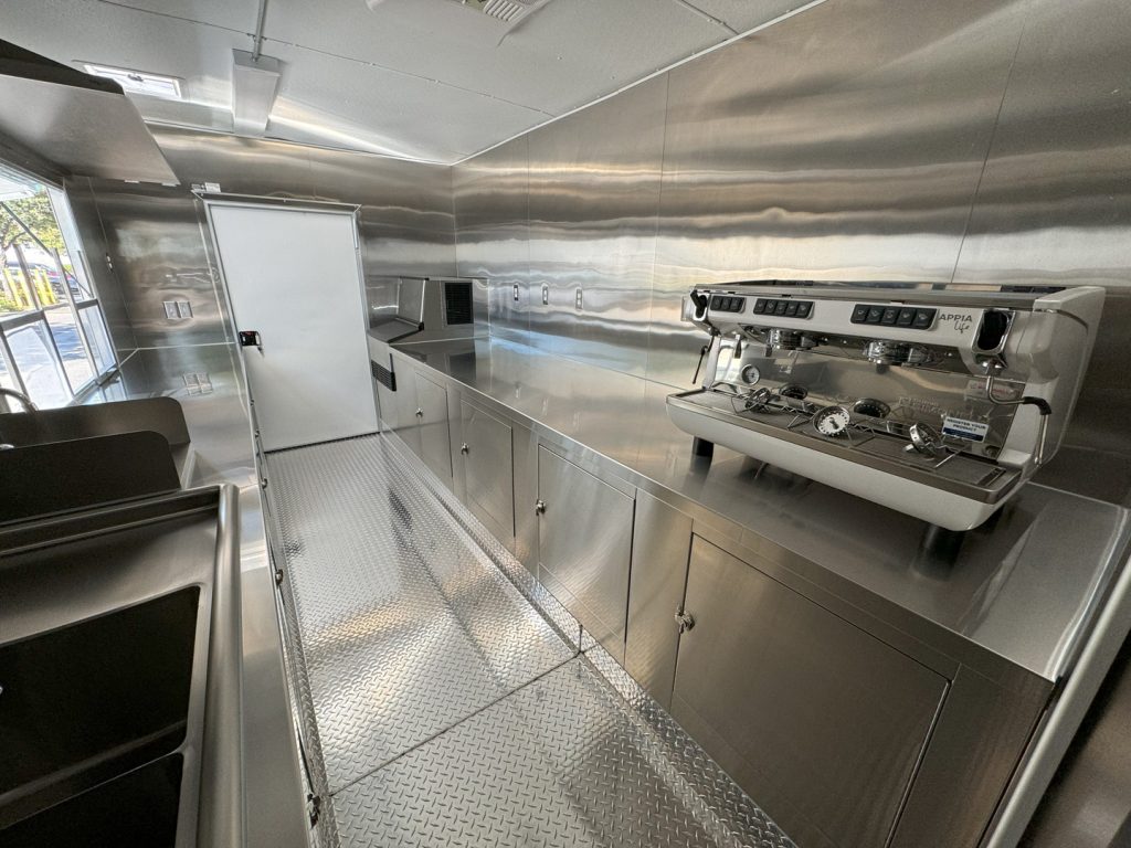 Commercial coffee trailer interior with an espresso machine, stainless steel counters, and beverage preparation area.