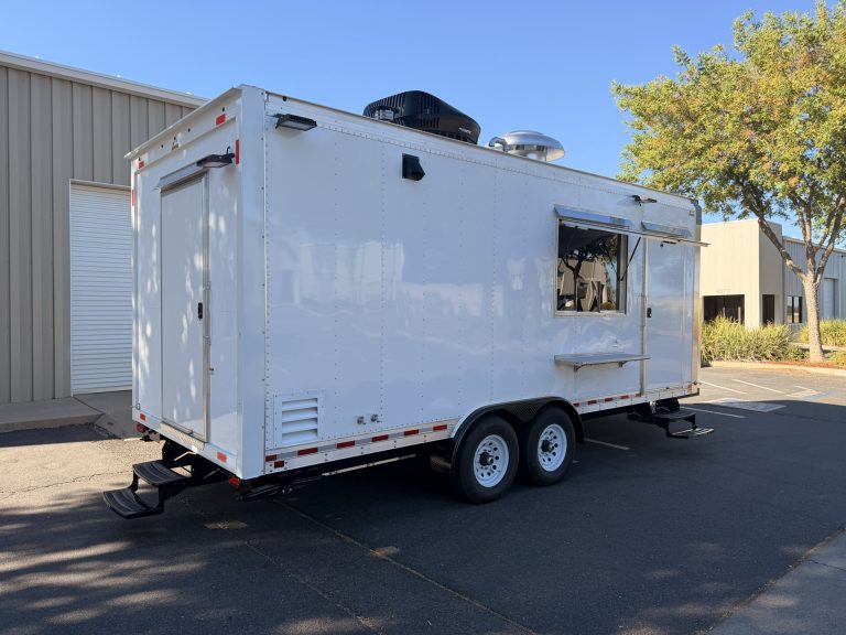 White custom commercial food trailer with serving windows, side entrance, and tandem axles parked outside the Golden State Trailers facility.
