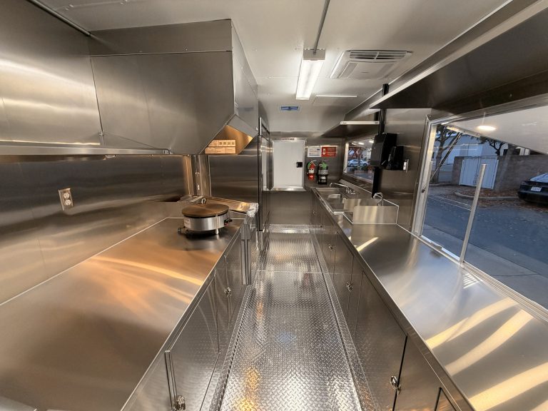 Commercial food trailer kitchen interior with stainless steel countertops, ventilation hood, equipment, and a central working aisle.