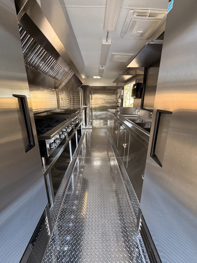 Custom food trailer interior with stainless steel cooking equipment, ventilation hood, sinks, refrigeration, and diamond-plate flooring.