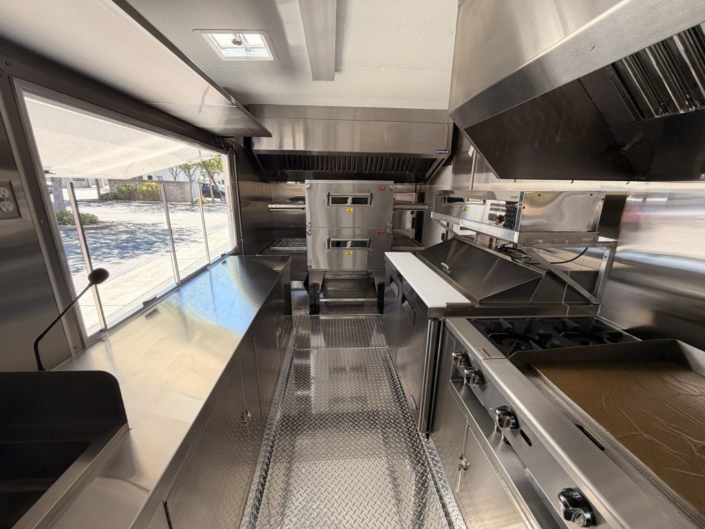 commercial pizza food trailer interior with service window, double-stack conveyor oven, hot plates, griddle, stainless steel counters, and ventilation hoods